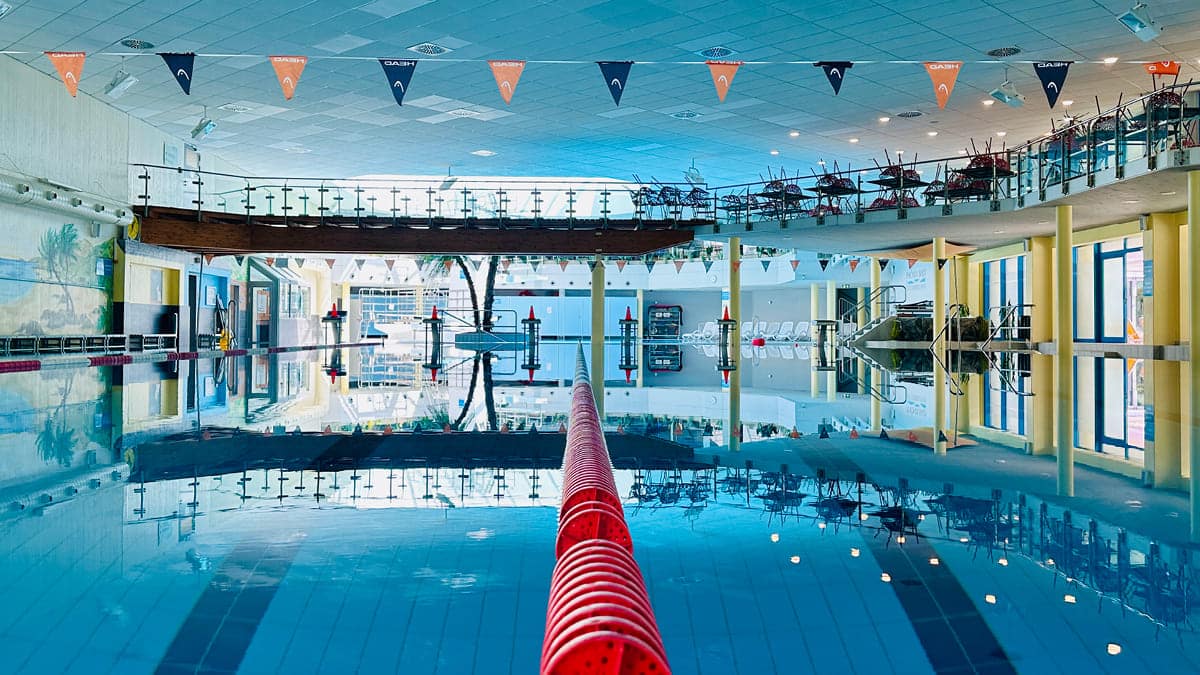 Blick in die leere Schwimmhalle, &uuml;ber das Schwimmerbecken im Ph&ouml;nix-Bad, der Mitte eine rote Trennleine zwischen den Schwimmbahnen; die Wasseroberfl&auml;che ist ganz still und die Umgebung spiegelt sich im Wasser